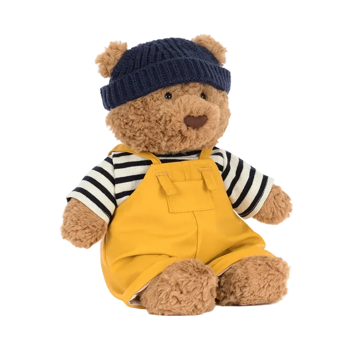 Teddy bear wearing a blue knitted hat, striped shirt, and yellow overalls on a white background