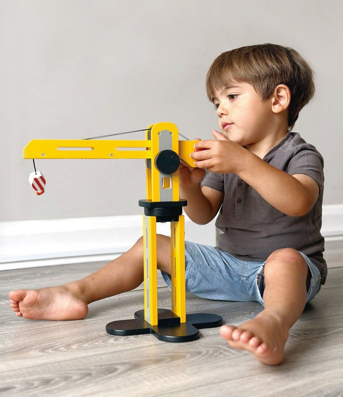 Young child sitting on the floor playing with a yellow wooden crane toy against a neutral background.