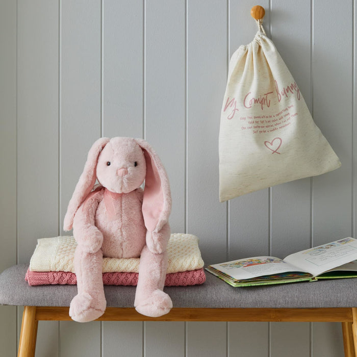Pink plush bunny, folded blanket, and open book on a bench with a drawstring bag hanging on the wall.